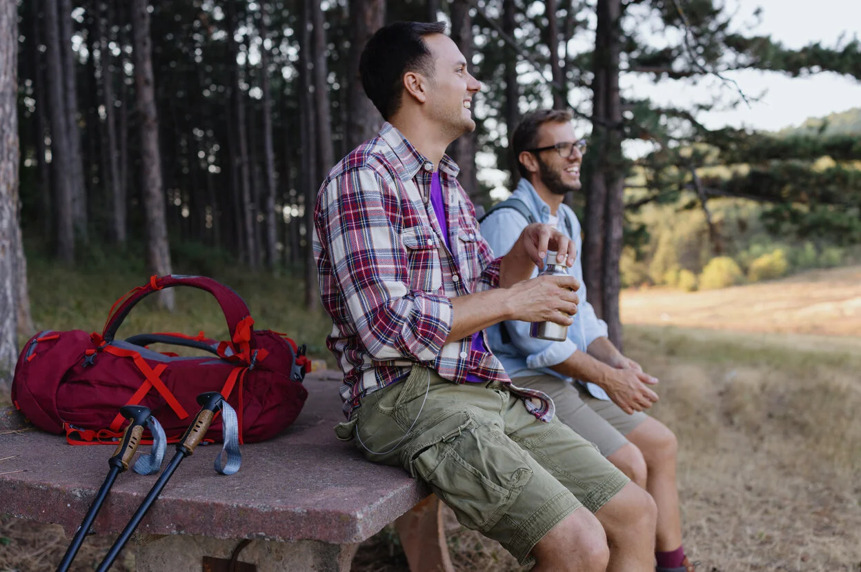 a couple of men sitting on a bench with a backpack and a bag